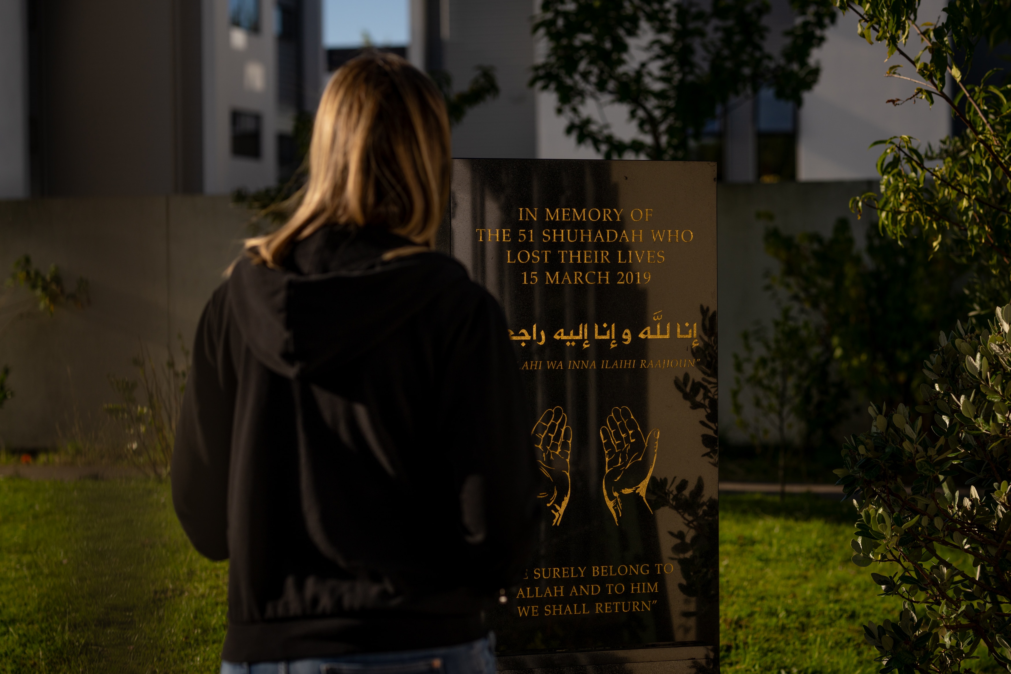 La espalda de una mujer con cabello rubio y un suéter negro parada frente a una placa con letras doradas.