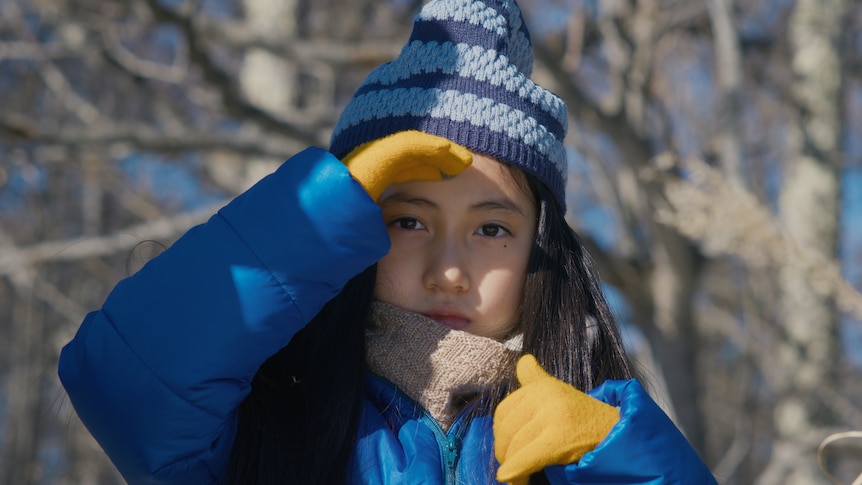 A young girl stares just beyond the camera, wearing a blue snow jacket and matching beanie. Her hands are in yellow gloves.