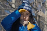 A young girl stares just beyond the camera, wearing a blue snow jacket and matching beanie. Her hands are in yellow gloves.