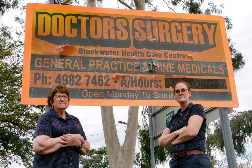 Two women standing with arms crossed in front of a sign.