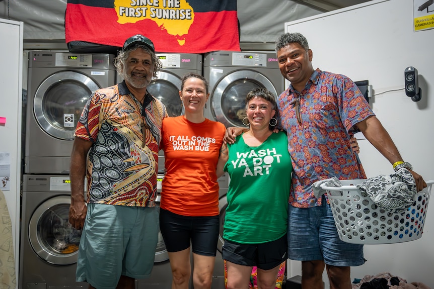 Four people stand smiling in front of washing machines.