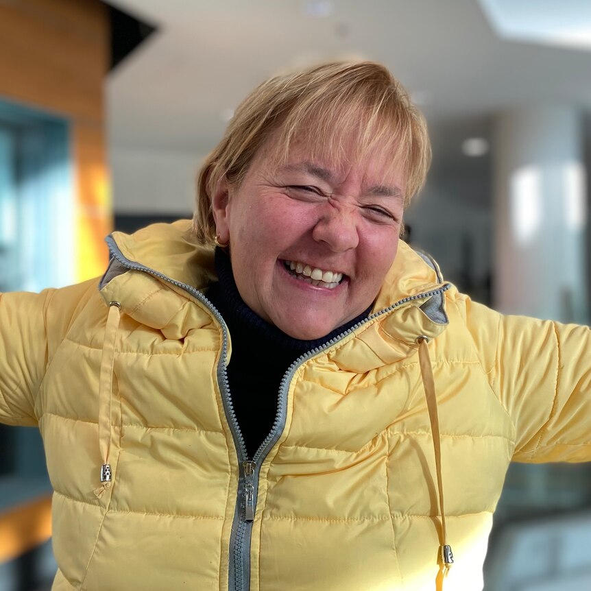 A woman with short blonde hair and a yellow parka grins with her arms spread wide in joy.