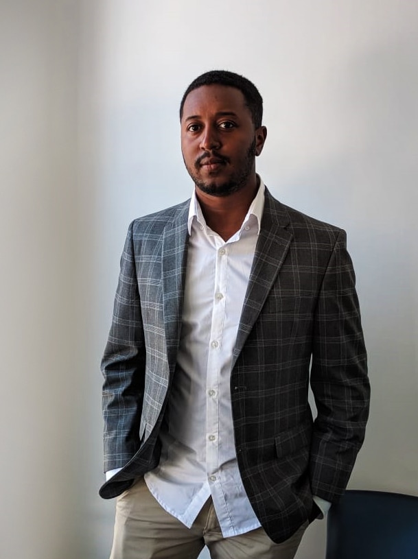 A man in a suit jacket stands against a wall as he poses for a photo.