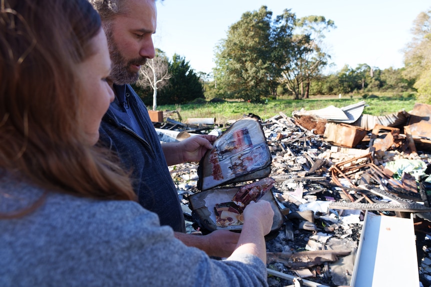 Justin and Verity stand in the ruins of their house looking at a burnt photo album on a sunny day.