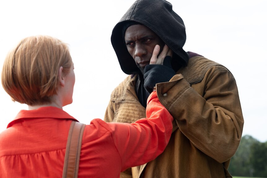 Woman in red top gently has her hand on face of taller Black man wearing a black hoodie and brown jacket. 