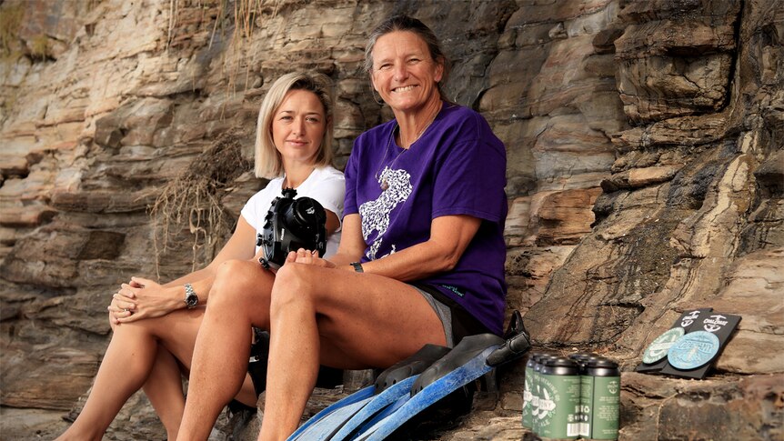 Kelly Carey and Lynne Tuck sit on rocks near the beach with a camera, flippers and beers in shot.