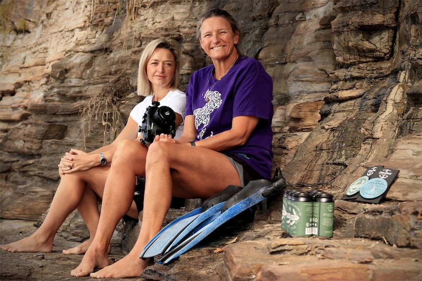 Kelly Carey and Lynne Tuck sit on rocks near the beach with a camera, flippers and beers in shot.