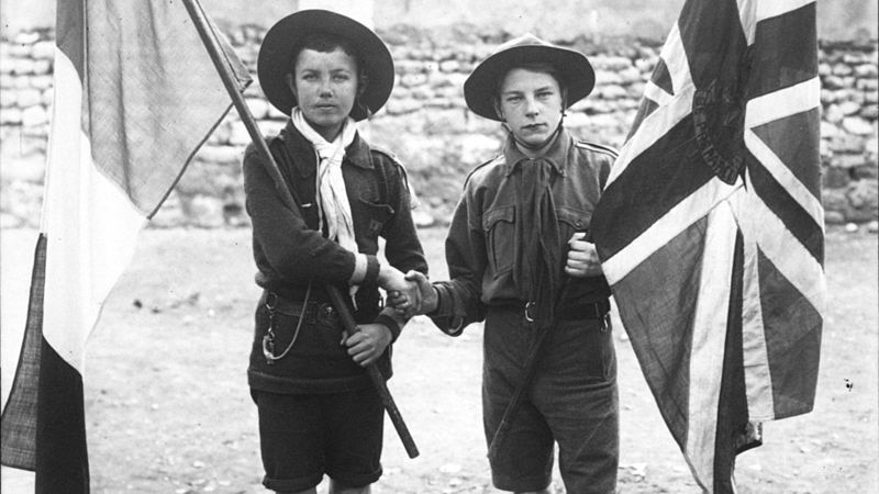 Two boys dressed in scouts uniform shaking hands while holding their national flags