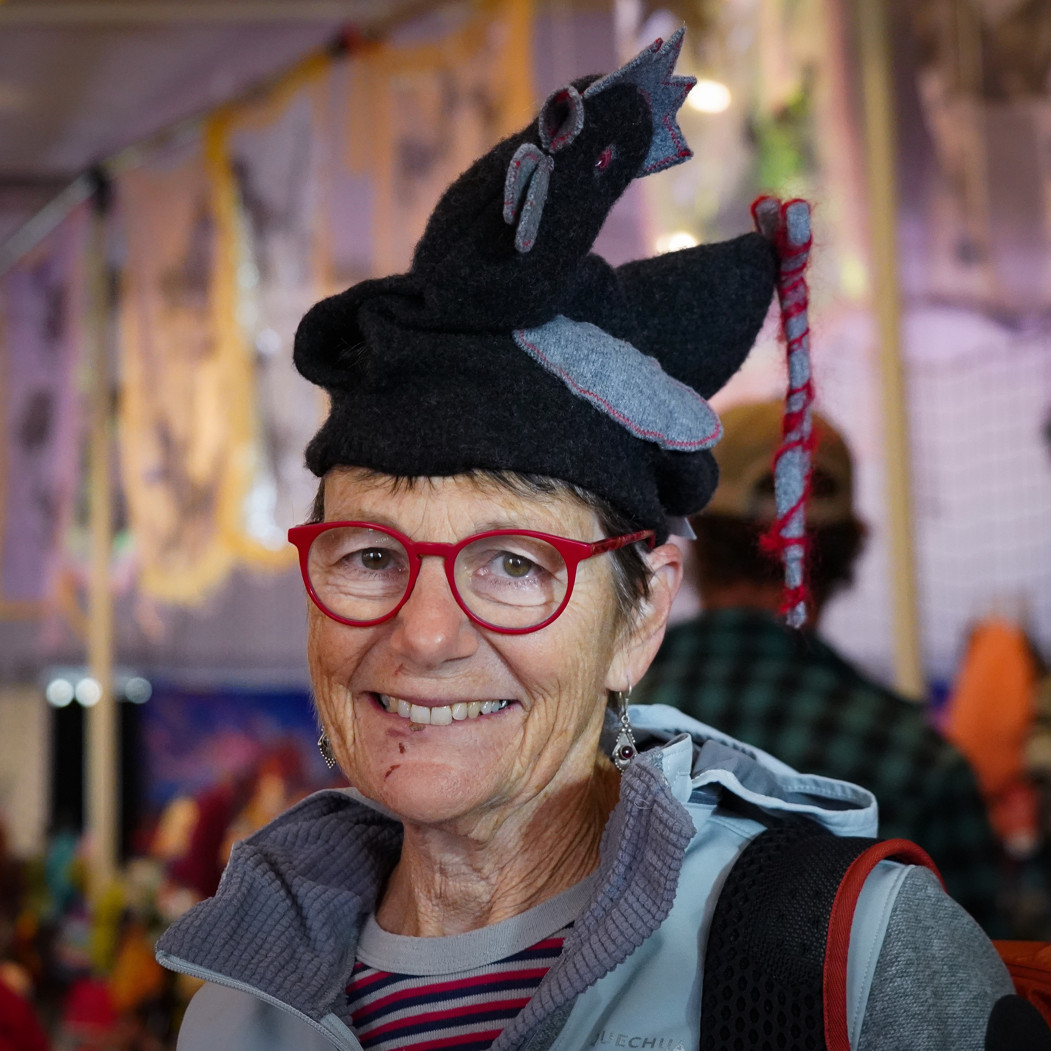 A woman smiles at the camera while wearing red-framed reading glasses and a beanie, knitted in the shape of a chicken.