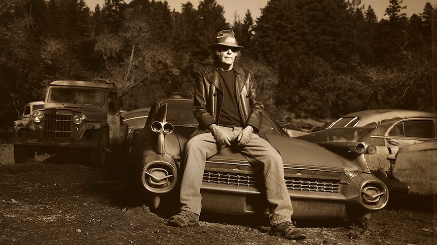Neil Young sits on the bonnet of an old vintage car in a junkyard. Other old cars sit behind him.