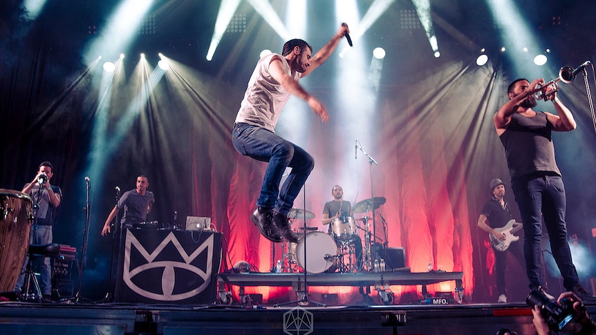 Lead singer of a band leaps into the air, while his band play on behind him 