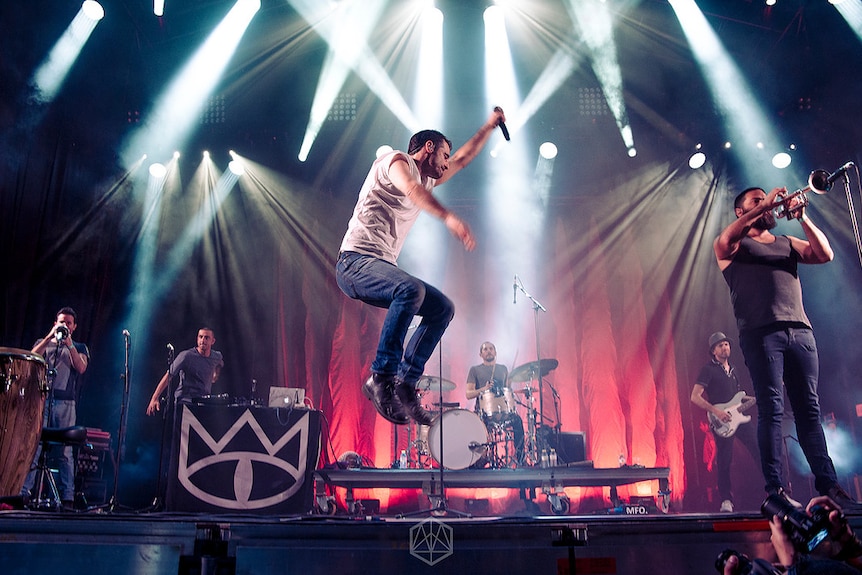 Lead singer of a band leaps into the air, while his band play on behind him 
