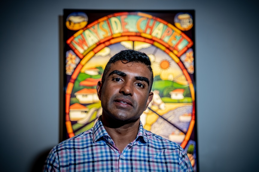 Jon Owen stands in front of a stained glass window, with his head tilted and a serious expression.