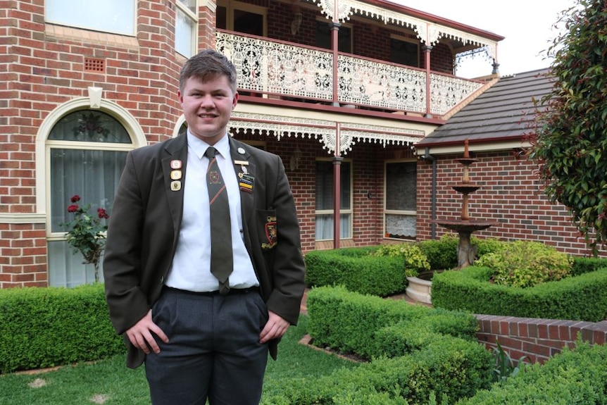 A young man in a school uniform stands in front of a house