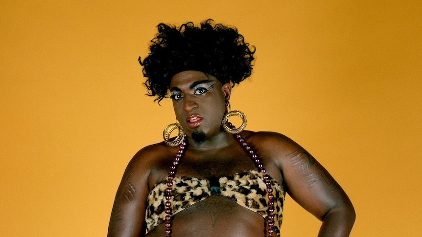 A tall black person with black hair and garish makeup wears high heels and a leopard print bikini against a yellow background