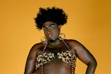 A tall black person with black hair and garish makeup wears high heels and a leopard print bikini against a yellow background