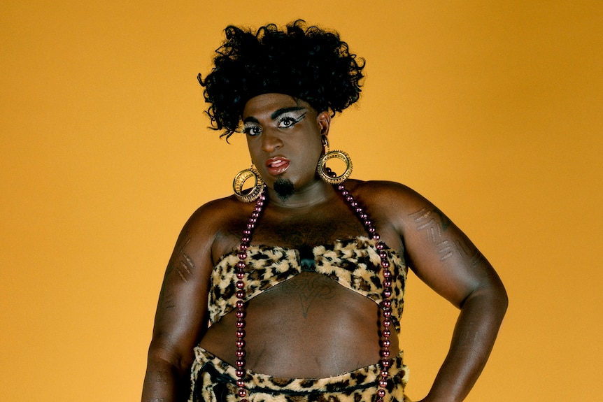 A tall black person with black hair and garish makeup wears high heels and a leopard print bikini against a yellow background