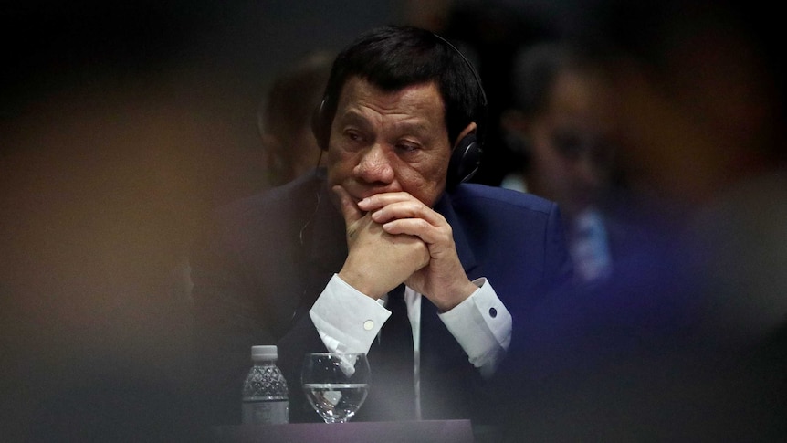 Rodrigo Duterte in focus in between two unidentified shoulders, rests his head on his hands. He is wearing headphones.