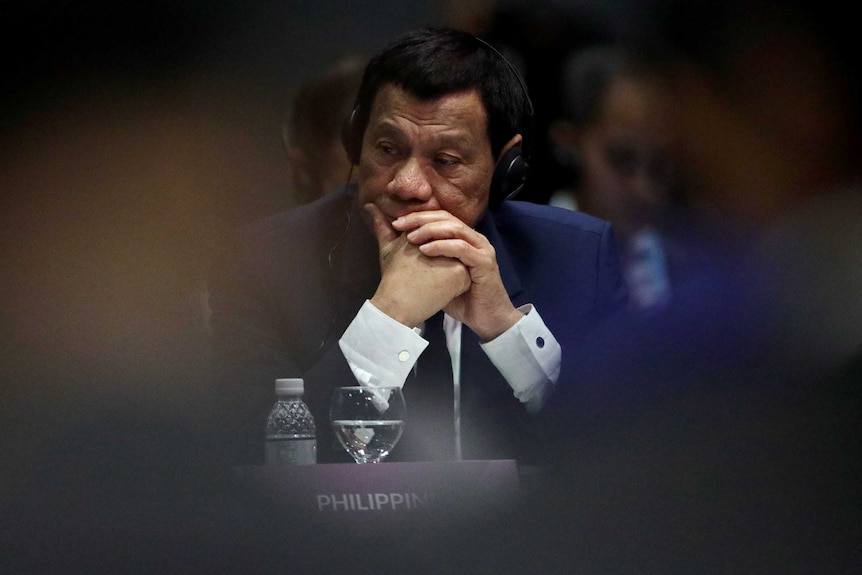 Rodrigo Duterte in focus in between two unidentified shoulders, rests his head on his hands. He is wearing headphones.