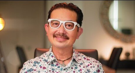 A Chinese man with white glasses and a flowery shirt smiles at the camera