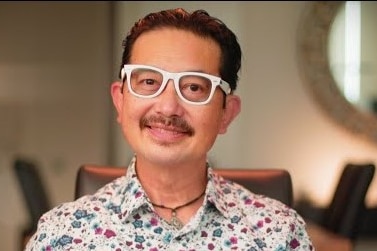A Chinese man with white glasses and a flowery shirt smiles at the camera