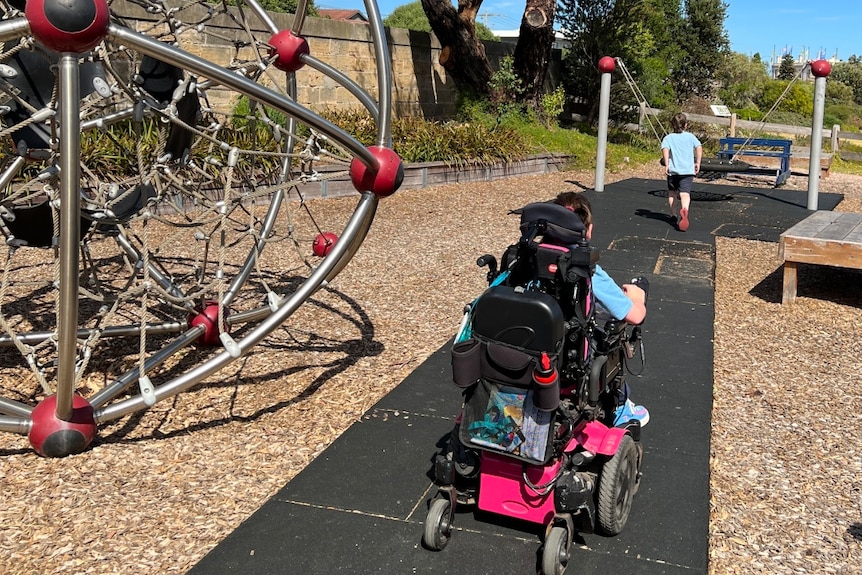 at the park, a boy runs over to the swing, his sister follows in a her wheelchair  