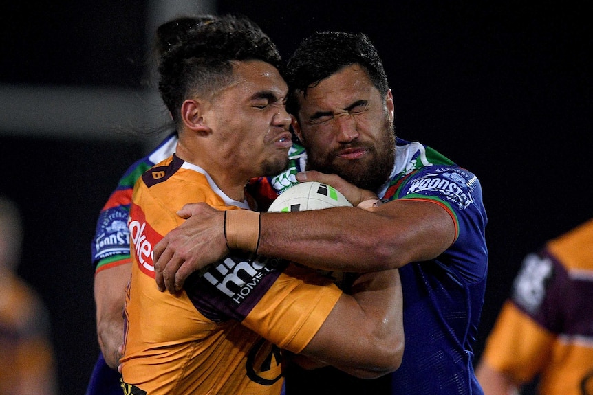 A Brisbane Broncos NRL player holding the ball is tackled across his upper body by a Warriors opponent.
