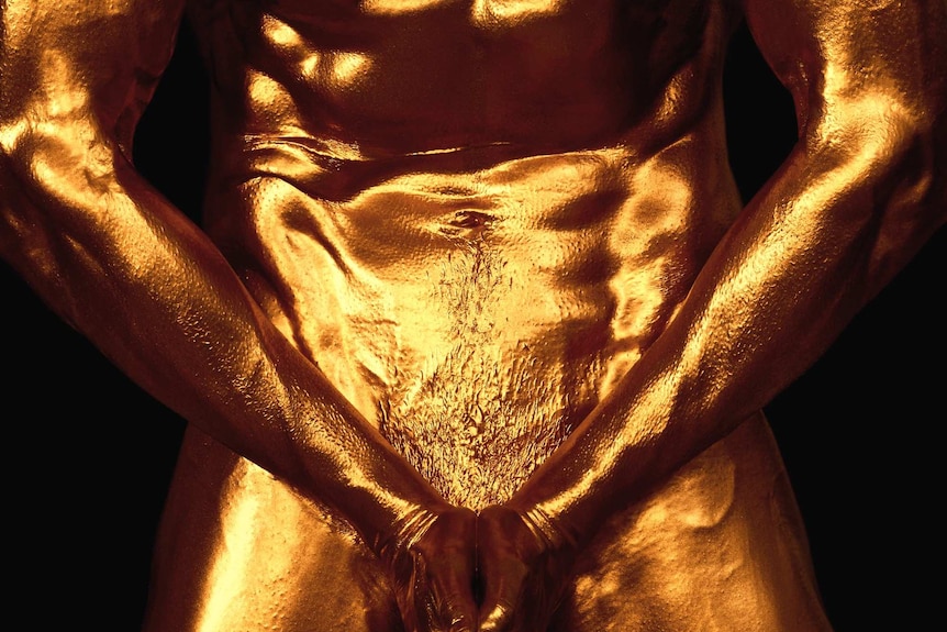 A naked muscled torso painted entirely in gold, pointing their hands downwards to hide their genitals.