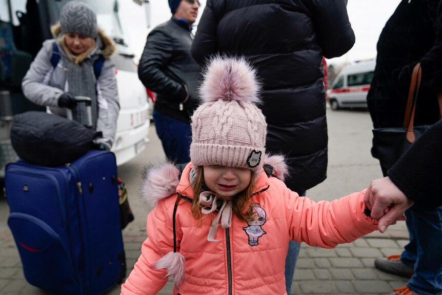 a with a beanie looks down as she is lead by the hand into Poland while other people stand behind her