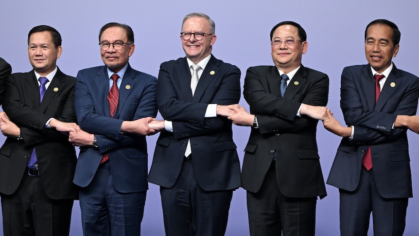 A wide shot of five men in suits and ties standing in a line, crossing their arms and joining hands while smiling