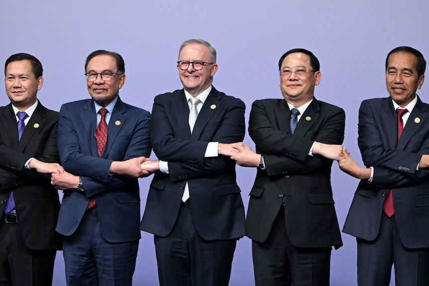 A wide shot of five men in suits and ties standing in a line, crossing their arms and joining hands while smiling