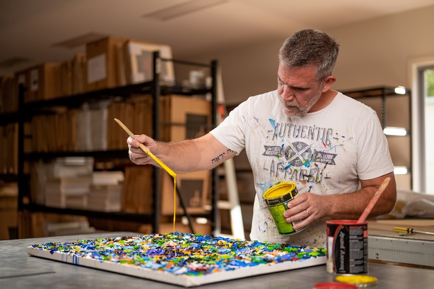 A man in his 60s in white tshirt with grey hair dabs paint onto a canvas