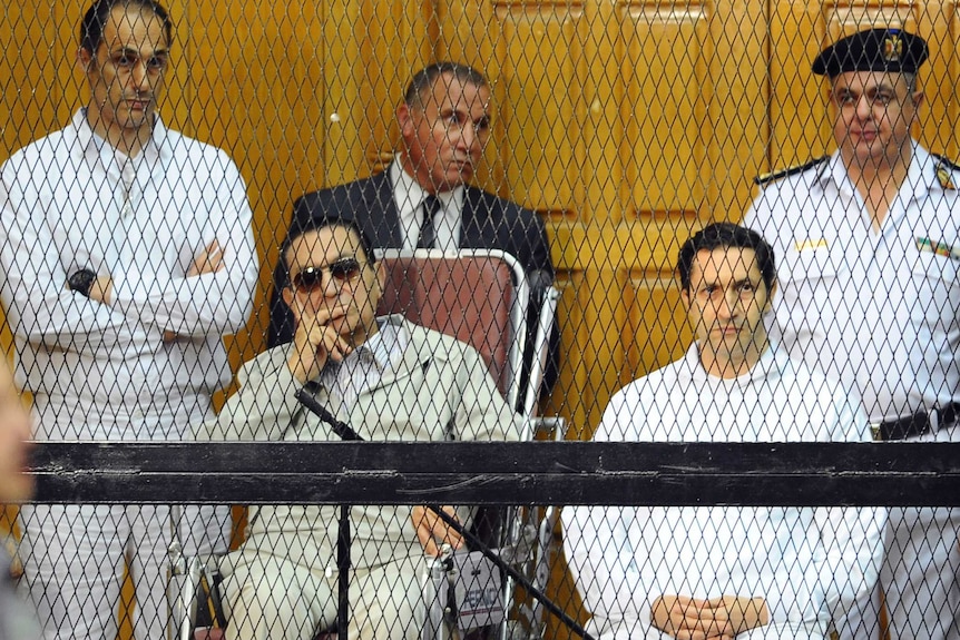 Hosni Mubarak, seated centre left, and his two sons, Gamal Mubarak, left, and Alaa Mubarak sit behind the fence in a courtroom.