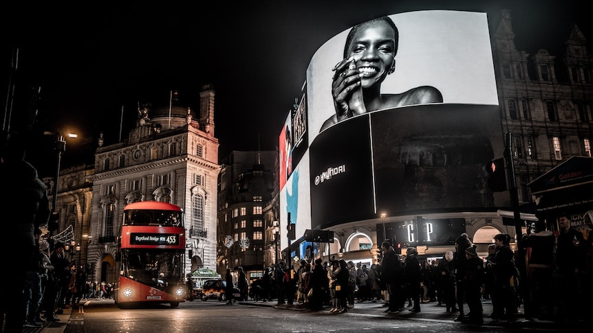 At night, you look up to large screens in London's Piccadilly Circus with a modern red double-decker pass driving towards you.