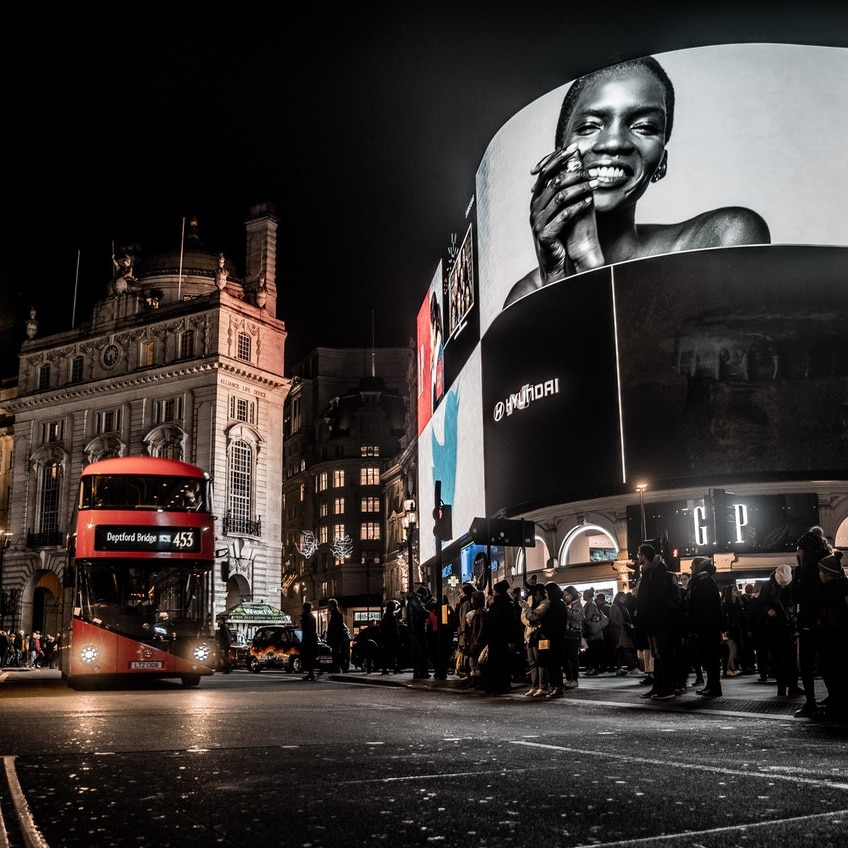 At night, you look up to large screens in London's Piccadilly Circus with a modern red double-decker pass driving towards you.