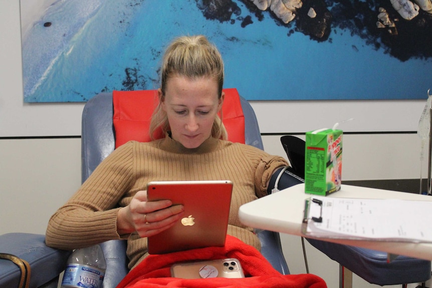 A woman with blonde hair wearing a brown jumper sits in the blood donation chair looking at an ipad with a juice box