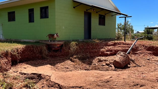 A cinder-block green-painted house, with a large dirt crater opened up beside it. A dog looks in from the edge.