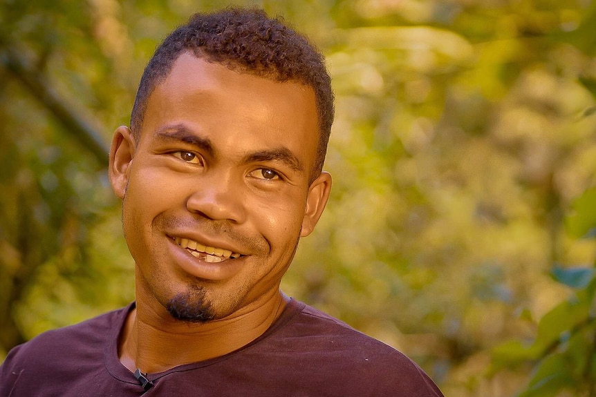 Mr Razafindramora looks to the right of the camera with a grin on his face. Plants are seen in the background.