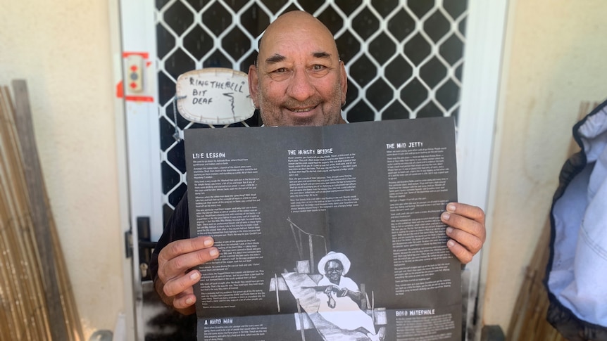 man holding large black fold-out pamphlet standing at front door. Sign on door made from lid says "Ring the bell. Bit deaf".