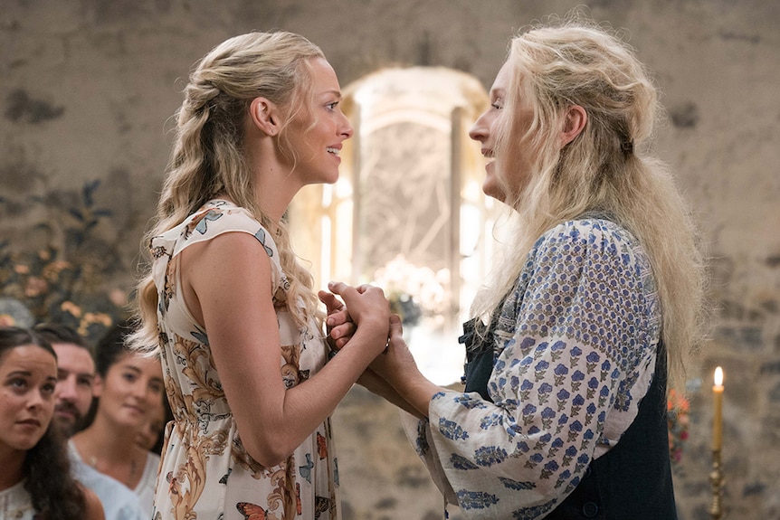 Colour image rom 2018 film of Amanda Seyfried and Meryl Streep clutching each other's hands in a rustic church chapel.