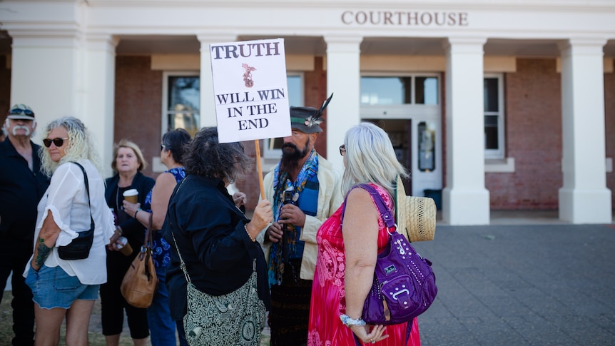 People speaking outside court with one holding a placard reading 'truth will win in the end'