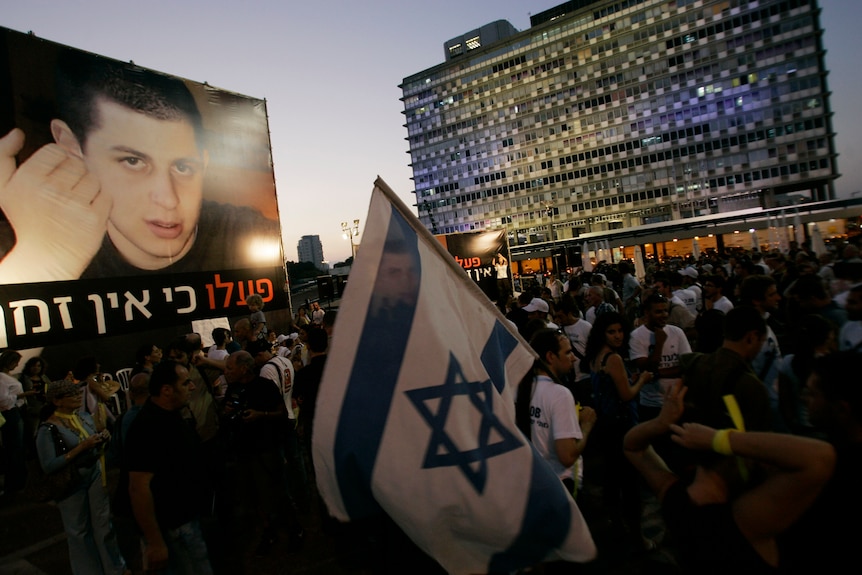 People wave Israeli flags and a poster of a young man's face, during a protest in an open city square