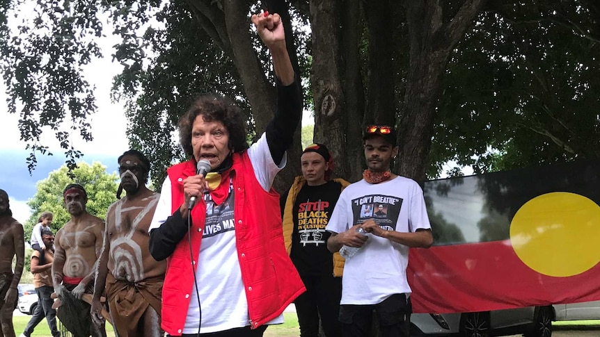 An older Indigenous woman, dressed in a red sleeveless jacket, raises her fist while addressing a crowd.