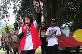 An older Indigenous woman, dressed in a red sleeveless jacket, raises her fist while addressing a crowd.