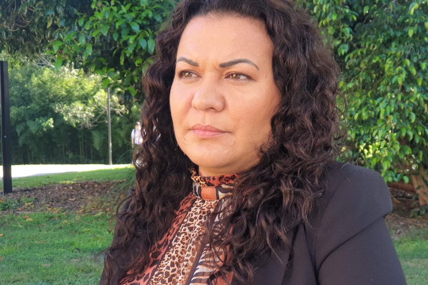 A headshot of a woman in a park. She is looking off-camera and has a neutral expression.