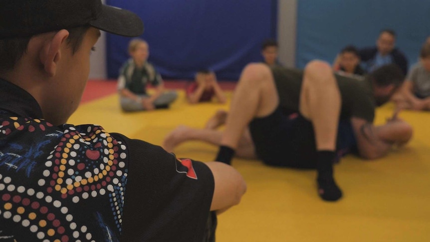The back of a young boy sitting on the floor watching a wrestling class.