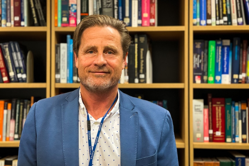 A man in a light blue suit and white spotted shirt in front of a bookshelf