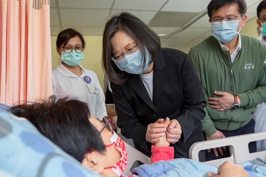 A woman leans over a hospital bed holding the patient's hand.