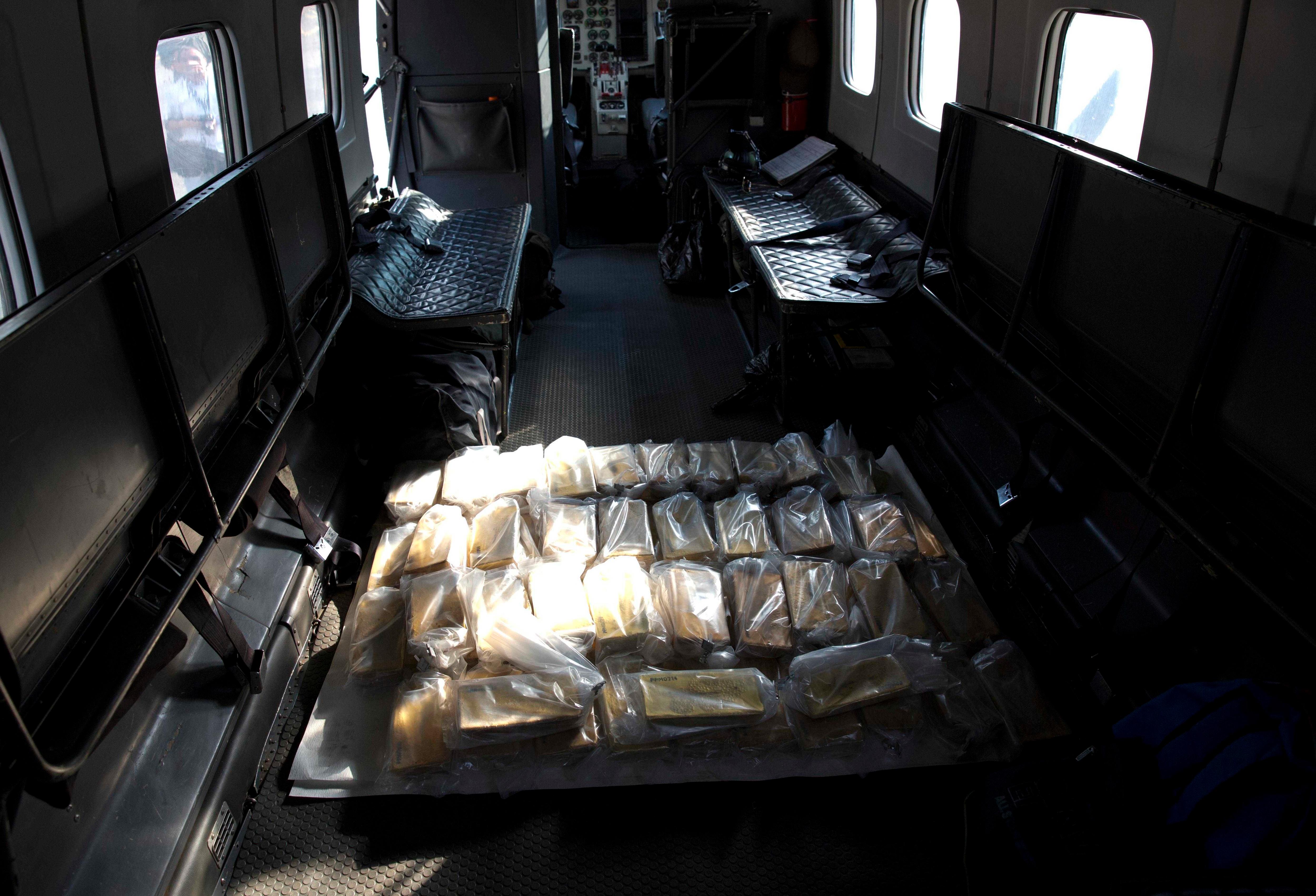 Gold bars wrapped in plastic sitting on a black platform inside a plane with black seats and open windows.