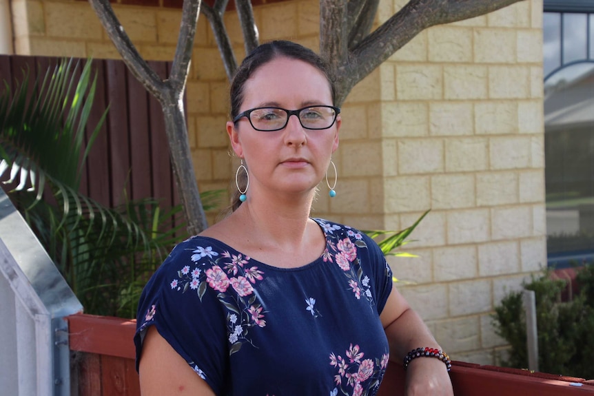 A mid-shot of woman wearing glasses faces the camera outside her home with a serious expression.